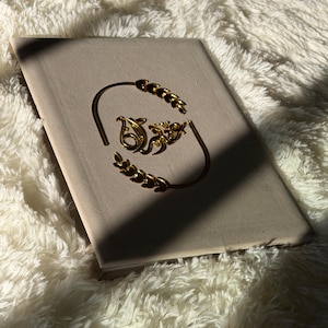 May include: A gold-colored bracelet with a decorative design, resting on a light-colored book. The bracelet features a curved design with leaf-like accents and an ornate central element. The background is a soft, white, fluffy texture.