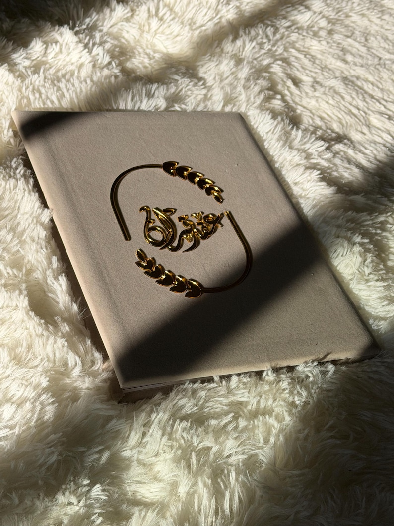 May include: A gold-colored bracelet with a decorative design, resting on a light-colored book. The bracelet features a curved design with leaf-like accents and an ornate central element. The background is a soft, white, fluffy texture.
