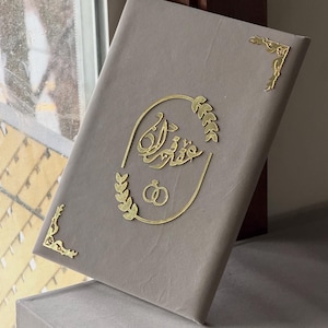 May include: A gray book with gold accents. The cover features gold script and leaf designs, along with two interlocking rings. Gold corner embellishments add detail. The book is resting on a gray surface.