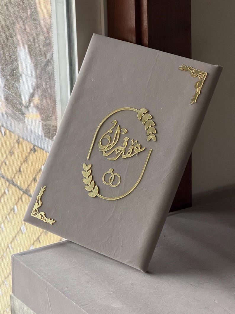 May include: A gray book with gold accents. The cover features gold script and leaf designs, along with two interlocking rings. Gold corner embellishments add detail. The book is resting on a gray surface.
