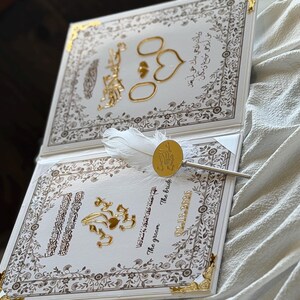 May include: A wedding invitation with gold accents and ornate floral borders. The invitation features gold rings, hearts, and decorative elements. A pen with a white feather and gold seal rests on the invitation. The date 02-15-2020 is visible.