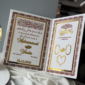 May include: A white wedding keepsake book with gold accents and Arabic calligraphy. The left side features names "Mohammed" and "Waela", the date 02.14.2026, and a heart design. The right side has a quote and ring and heart illustrations.