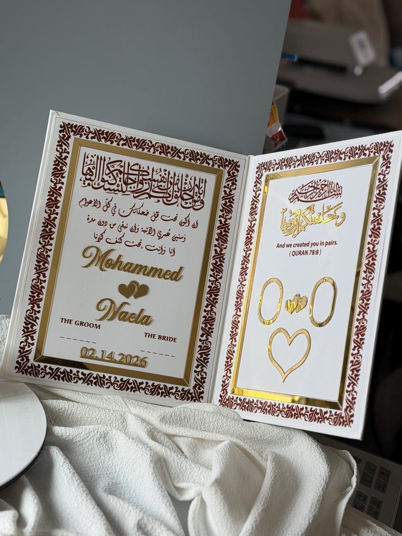 May include: A white wedding keepsake book with gold accents and Arabic calligraphy. The left side features names "Mohammed" and "Waela", the date 02.14.2026, and a heart design. The right side has a quote and ring and heart illustrations.