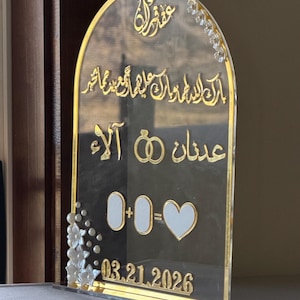 May include: Clear acrylic wedding sign with gold accents. The sign features Arabic calligraphy, two gold rings, and the date 03.21.2026. White floral embellishments adorn the left side and top right corner. The sign is designed to stand upright.