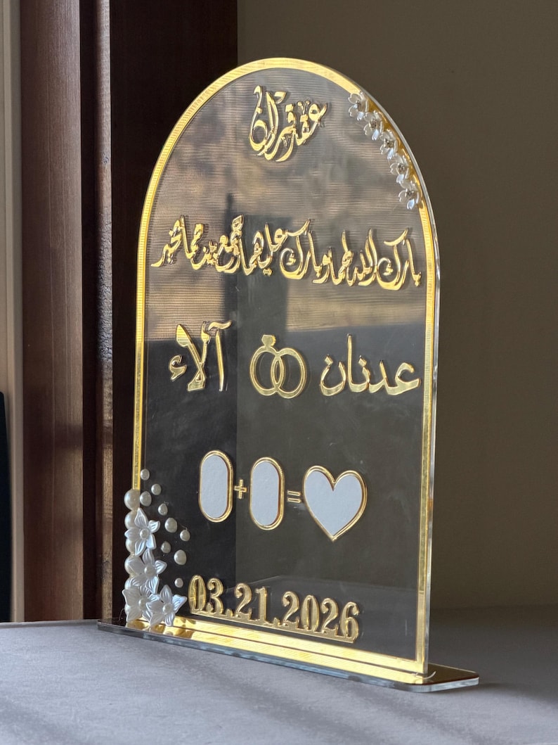 May include: Clear acrylic wedding sign with gold accents. The sign features Arabic calligraphy, two gold rings, and the date 03.21.2026. White floral embellishments adorn the left side and top right corner. The sign is designed to stand upright.
