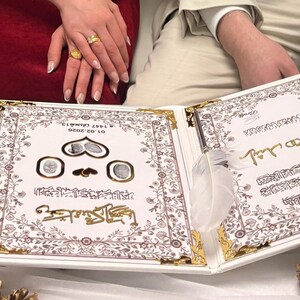 May include: An open, white wedding album with gold accents and ornate floral designs. The album features Arabic script and decorative elements. Gold-colored pine cones and a compact mirror are also visible.