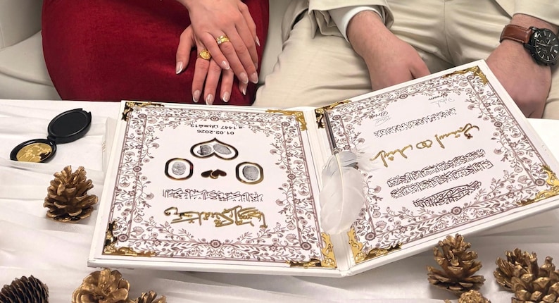May include: An open, white wedding album with gold accents and ornate floral designs. The album features Arabic script and decorative elements. Gold-colored pine cones and a compact mirror are also visible.