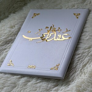 May include: A white book with gold accents and Arabic script. The cover features gold corner designs and the year 2026. The Arabic script is in a gold color and is the focal point of the book's design.