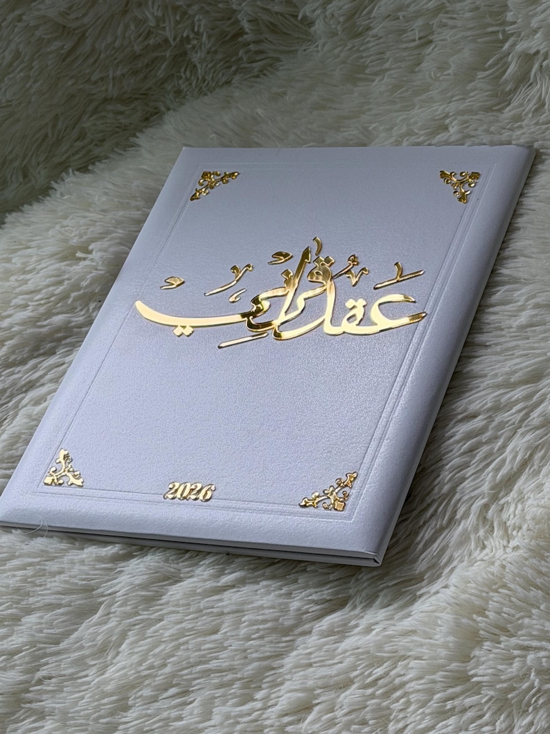 May include: A white book with gold accents and Arabic script. The cover features gold corner designs and the year 2026. The Arabic script is in a gold color and is the focal point of the book's design.