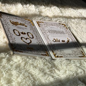 May include: Open book with gold accents and Arabic script. The pages are white with gold borders and feature gold rings and a heart. The book is on a fluffy white surface, with sunlight casting shadows.