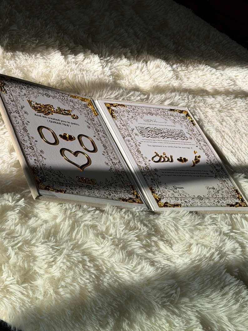 May include: Open book with gold accents and Arabic script. The pages are white with gold borders and feature gold rings and a heart. The book is on a fluffy white surface, with sunlight casting shadows.