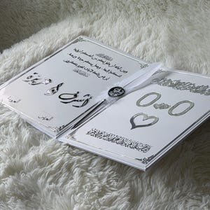 May include: Two white wedding invitation cards with silver accents and Arabic script. The cards are open, revealing text and decorative elements, including hearts and rings. A white ribbon and a decorative button are attached.