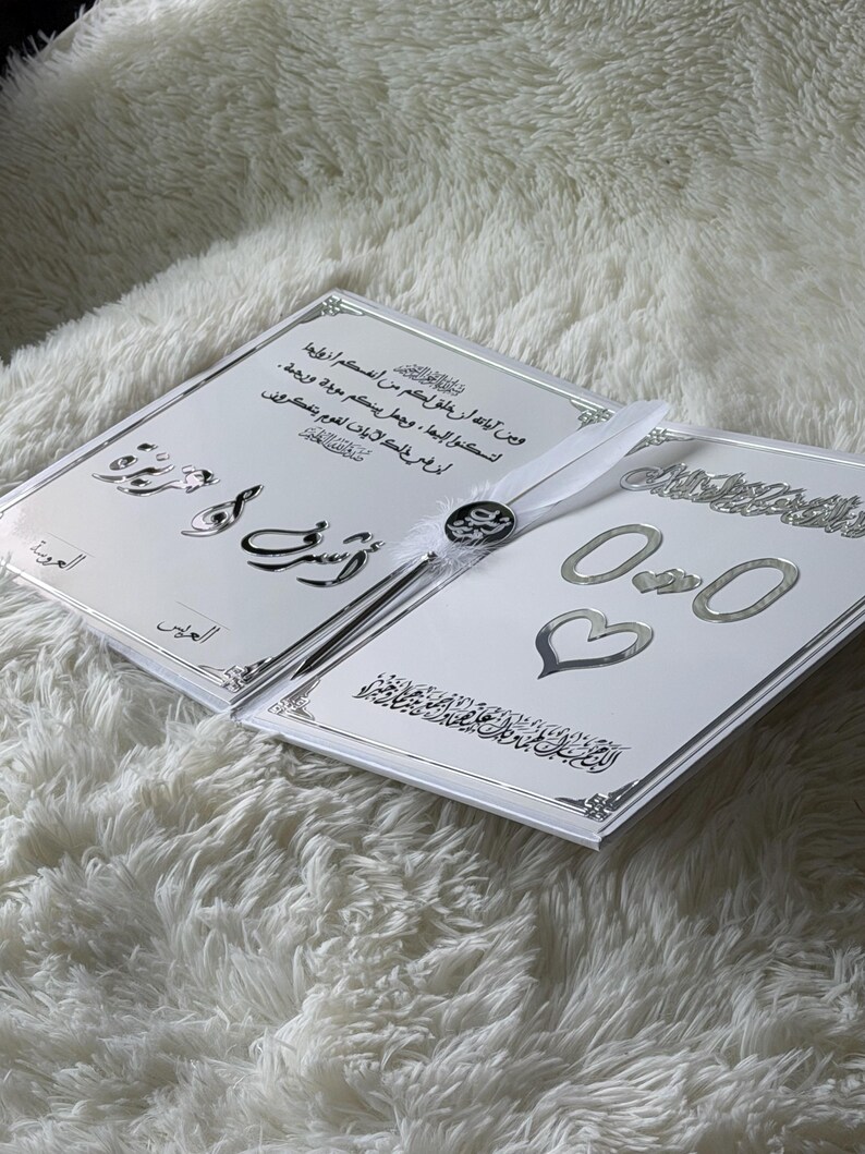 May include: Two white wedding invitation cards with silver accents and Arabic script. The cards are open, revealing text and decorative elements, including hearts and rings. A white ribbon and a decorative button are attached.