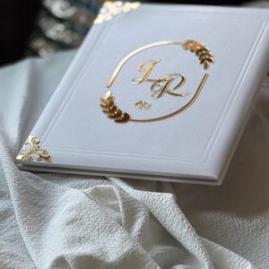 May include: A white photo album with gold accents. The cover features gold initials inside a gold oval with wheat-like designs. Gold corner protectors add a decorative touch. The album rests on a white textured fabric.