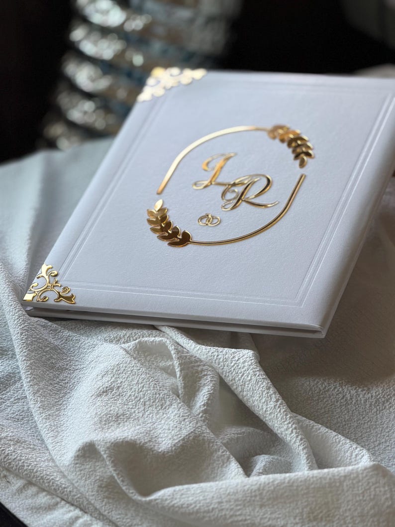 May include: A white photo album with gold accents. The cover features gold initials inside a gold oval with wheat-like designs. Gold corner protectors add a decorative touch. The album rests on a white textured fabric.