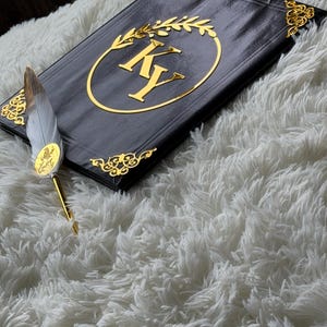 May include: Black journal with gold accents, featuring the initials "KY" inside a gold laurel wreath. The journal has gold corner embellishments and is paired with a feather pen with gold details. The items rest on a fluffy white surface.