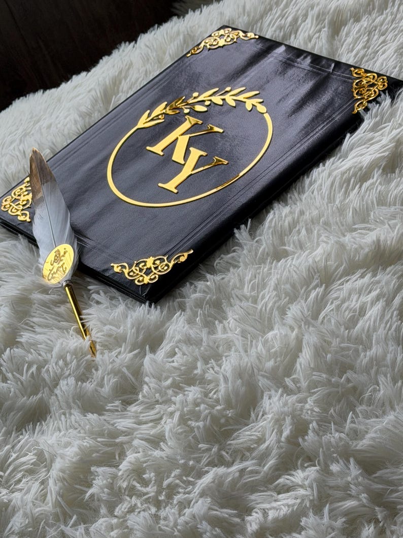 May include: Black journal with gold accents, featuring the initials "KY" inside a gold laurel wreath. The journal has gold corner embellishments and is paired with a feather pen with gold details. The items rest on a fluffy white surface.