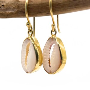 Könnte beinhalten: Ein Paar goldfarbene Ohrhänger mit einem natürlichen Muscheldesign. Die Muscheln sind in einen goldfarbenen Metallrahmen eingefasst, mit einem Hakenverschluss. Die Ohrringe werden auf einem Holzast vor weißem Hintergrund präsentiert.
