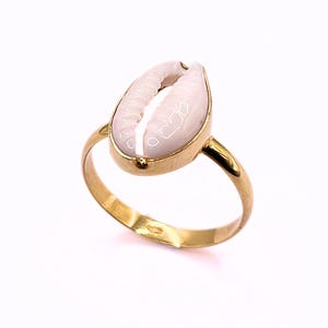 Könnte beinhalten: Goldener Ring mit einer weißen Muschel. Der Ring hat ein schlichtes Band und eine Fassung, die die Muschel hält. Die Muschel ist oval und hat eine strukturierte Oberfläche.