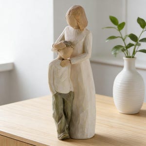 May include: A carved figurine of a mother and child. The mother figure is in a long, flowing dress, with her arm around the child. The child is wearing a shirt and pants. The color palette is neutral, with shades of beige and green. The figurine is on a wooden surface.