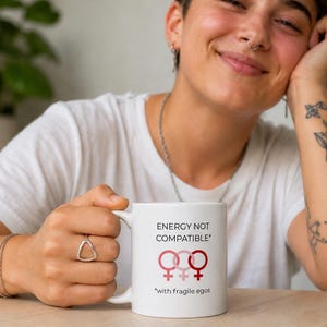 Könnte beinhalten: Ein weißer Keramikbecher mit dem schwarzen Text "ENERGY NOT COMPATIBLE" über drei rosa und roten weiblichen Geschlechtssymbolen sowie dem zusätzlichen schwarzen Text "*with fragile egos" auf der unteren Außenseite des Bechers.