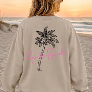 Könnte beinhalten: Ein beigefarbenes Sweatshirt mit Rundhalsausschnitt und einer schwarzen Strichzeichnung einer Palme auf der Rückseite. Über den Stamm des Baumes ist der Text "Rose & Respek!" in einer leuchtend pinkfarbenen Schreibschrift gedruckt.