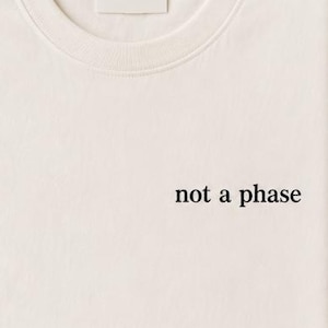 Peut inclure: T-shirt crème plié avec l'inscription noire "not a phase". Le t-shirt est posé sur une surface neutre, avec une branche de verdure et une couverture en tricot beige en arrière-plan. Le t-shirt a une étiquette vierge.