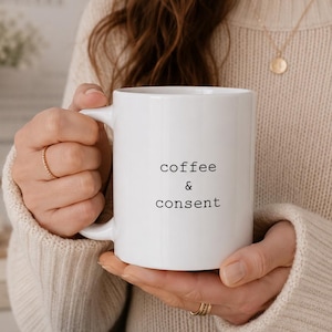 Op de afbeelding: Witte keramische mok met de woorden "coffee & consent" in zwarte letters. De mok heeft een gebogen oor en wordt vastgehouden door een persoon die een crèmekleurige trui draagt.