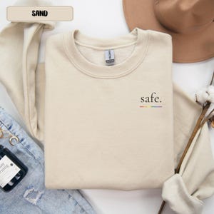 Könnte beinhalten: Ein sandfarbenes Sweatshirt mit Rundhalsausschnitt, auf dessen linker Brustseite das Wort "safe." in Schwarz gedruckt ist, mit einer dünnen horizontalen regenbogenfarbenen Linie direkt unter dem Text. Das Kleidungsstück hat lange Ärmel und einen Rippkragen.