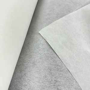 May include: Close-up of white, non-woven fabric in a roll and a flat sheet. The fabric has a soft, slightly textured appearance. The background is a light grey, creating a contrast with the white fabric. The image highlights the texture and colour of the material.