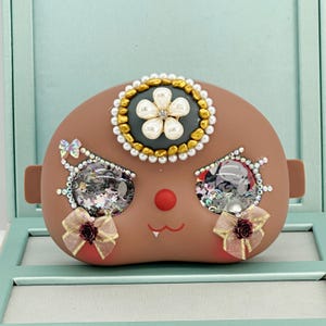 May include: A brown, face-shaped decorative object with a flower design on top, embellished with pearls and rhinestones. The eyes are filled with sparkling elements, and bows adorn the cheeks. A red nose and a smiling mouth complete the design.