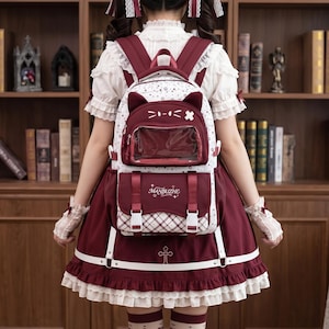 Könnte beinhalten: Ein bordeauxroter und weißer Rucksack mit Katzenohren-Design, einer transparenten Vordertasche und einer unteren Klappe mit dem Schriftzug MANBUZHE. Der Rucksack verfügt über verstellbare Träger, Schnallenverschlüsse und ein Sternenmuster.