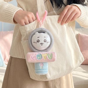 May include: A plush toy keychain featuring a white cartoon character with a round face inside a clear dome, attached to a pink coiled strap. Below the dome is a rectangular pink plush bar with the letters COTV and a light blue handle base.