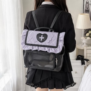 Könnte beinhalten: Ein schwarz-helllila Rucksack mit einer gesteppten Klappe mit Rüschenrand und einem schwarzen Herzaufnäher in der Mitte, der ein weißes Kreuzsymbol zeigt. Der untere Teil enthält ein durchsichtiges rechteckiges Sichtfenster und eine Seitentasche.