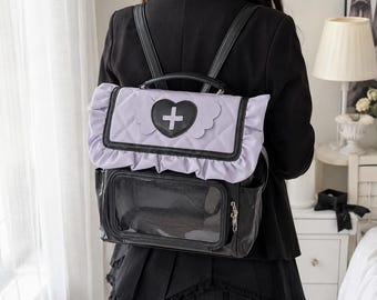 May include: A black and light purple backpack featuring a quilted flap with a ruffled edge and a central black heart patch containing a white cross symbol. The lower section includes a clear rectangular display window and a side zipper pocket.