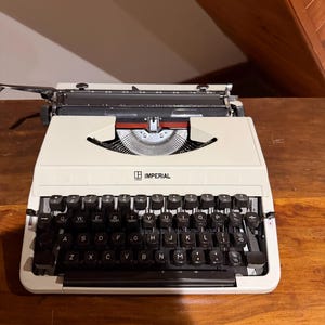 May include: Cream-coloured Imperial typewriter with black keys and a red accent. The typewriter sits on a wooden surface. The word "IMPERIAL" is printed on the front of the typewriter.