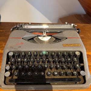 May include: A vintage gray typewriter with black keys and a red accent. The keyboard features QWERTY layout with round keys. The typewriter has a metal body and is placed on a wooden surface.