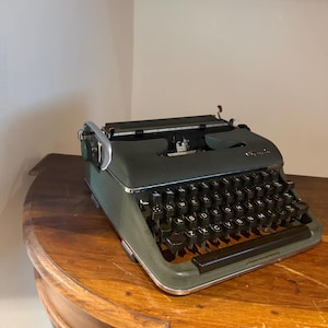 Peut inclure: Une machine à écrire manuelle Olympia vert foncé dotée d'un clavier QWERTY avec des touches circulaires noires, un levier de retour de chariot argenté et un cadre en métal. La marque Olympia est imprimée en lettres argentées sur le capot.