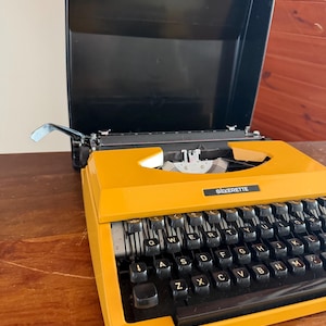 Puede incluir: Una máquina de escribir SILVERETTE amarilla vintage con teclas negras y una cubierta negra. La máquina de escribir está sobre una superficie de madera. Las teclas están dispuestas en un diseño QWERTY.