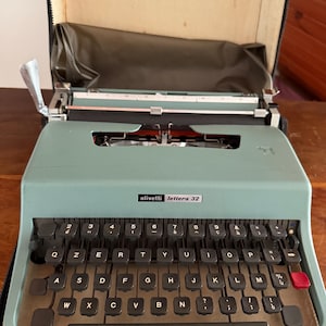 May include: A seafoam green Olivetti Lettera 32 typewriter with black keys and white lettering. The typewriter has a red return key. The machine is in its original carrying case. The case is open.
