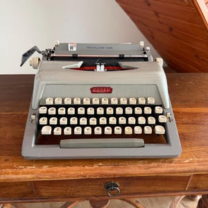 May include: A vintage Royaluxe 425 typewriter in grey with white keys. The typewriter sits on a wooden table. The keys are arranged in a QWERTY layout. The word "ROYAL" is displayed on the front of the typewriter.