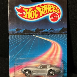 May include: A silver die-cast metal Hot Wheels car in its original packaging. The card features the Hot Wheels logo, the Mattel logo, and a retro design with the text "FOR AGES OVER 3. DIE CAST METAL!". The car is a classic sports car.