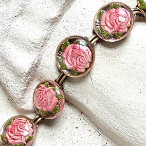 Może przedstawiać: Bransoletka w stylu vintage z owalnymi zawieszkami, z różowymi wzorami róż zamkniętymi w przezroczystej żywicy. Bransoletka ma łańcuszek i zapięcie z metalu w odcieniu złota. Róże mają zielone liście. Na teksturowanej białej powierzchni.