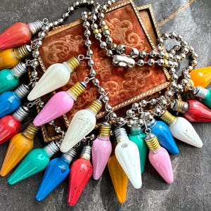 May include: A necklace featuring colorful, vintage-style Christmas light bulb pendants. The bulbs are in various colors, including red, yellow, green, blue, white, and pink. The necklace has a silver chain and a decorative, ornate, brown velvet box in the background.