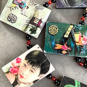 May include: A necklace with black and red beads and small rectangular pendants with Asian-inspired imagery. The pendants feature illustrations of people, buildings, and landscapes.
