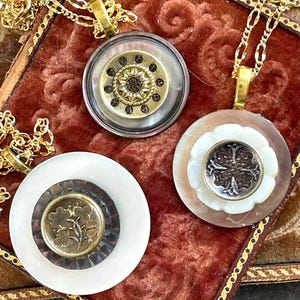 May include: Three vintage-style necklaces with gold chains and circular pendants. The pendants feature mother-of-pearl, brass, and decorative buttons. One pendant has a floral design, another has a sunburst pattern, and the third has a four-leaf clover.