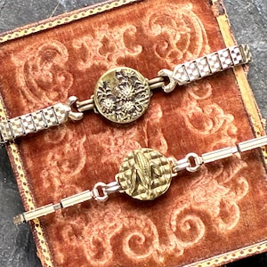 May include: Two vintage metal bracelets with decorative button clasps. One clasp features a floral design, the other a textured pattern. The bracelets are silver-toned with gold-toned clasps, set against a patterned, reddish-brown velvet surface.