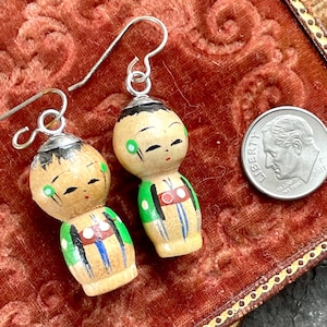 以下が含まれることがあります： 木製のヴィンテージ人形のイヤリングのペア。各イヤリングは、黒いトップス、緑と赤のアクセント、銀色のイヤーフックを備えた手描きの人形が特徴です。 イヤリングは赤いベルベットの表面にあります。