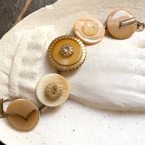 May include: A bracelet made of five round buttons with a gold trim. The buttons are beige and brown with a gold centre. The bracelet is on a white surface.