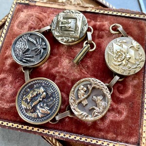May include: A vintage bracelet featuring six circular, ornate buttons with various relief designs. The buttons are connected by small metal links and are set against a red velvet background. The buttons have gold and silver tones.